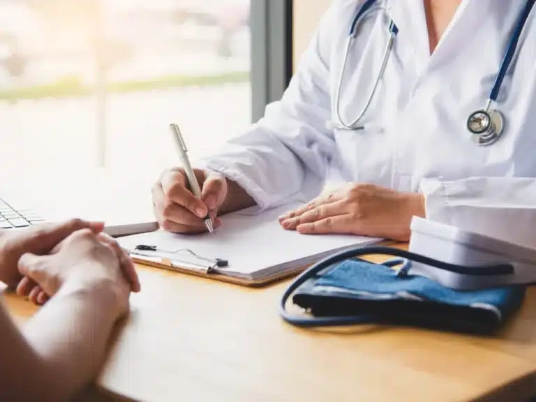 Doctor writing notes at her desk while a patient describes how they feel