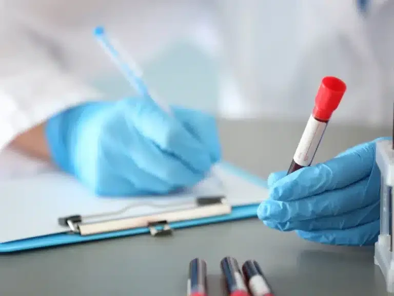 Laboratory technician in blue gloves holding a blood sample tube beside a form